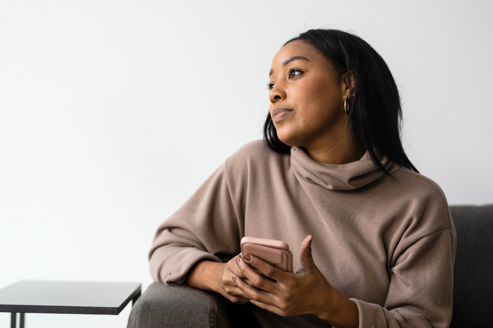 A bored woman stares off into the distance to her right. She is sitting in a plush charcoal grey chair, holding her phone in her hands.
