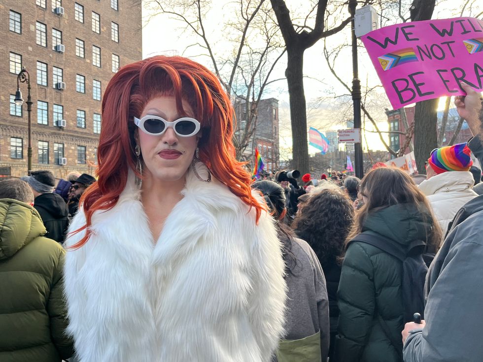 A drag queen with a red wig, round white glasses and a shaggy white fur coat looks at the camera with a solemn expression. Around her, a crowd of people look at something in the distance, many holding signs and rainbow-colored LGBTQ Pride paraphernalia.