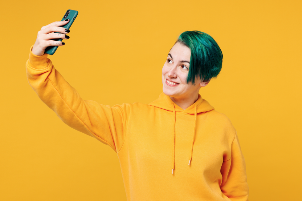A picture of a woman with short green hair in a yellow hoodie against a yellow background taking a selfie.\u200b