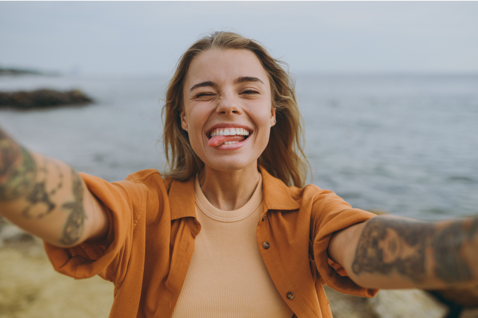 A selfie of a woman standing by the ocean. She is smiling while sticking her tongue out and scrunching her face slightly.