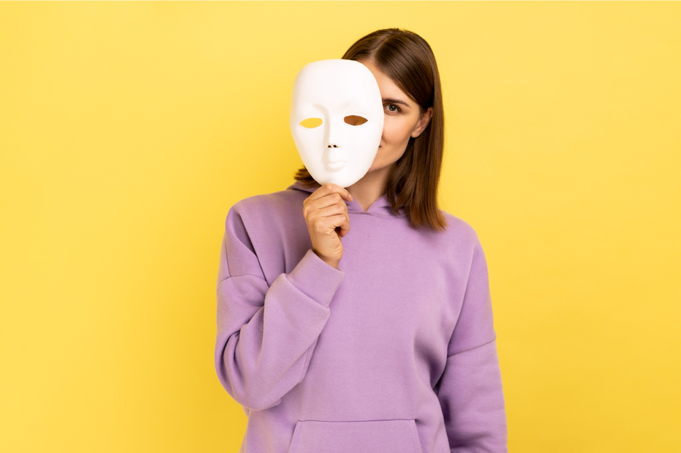 A woman in a purple hoodie in front of a yellow background. She is peeking out from behind a white mask that she's holding at the chin.