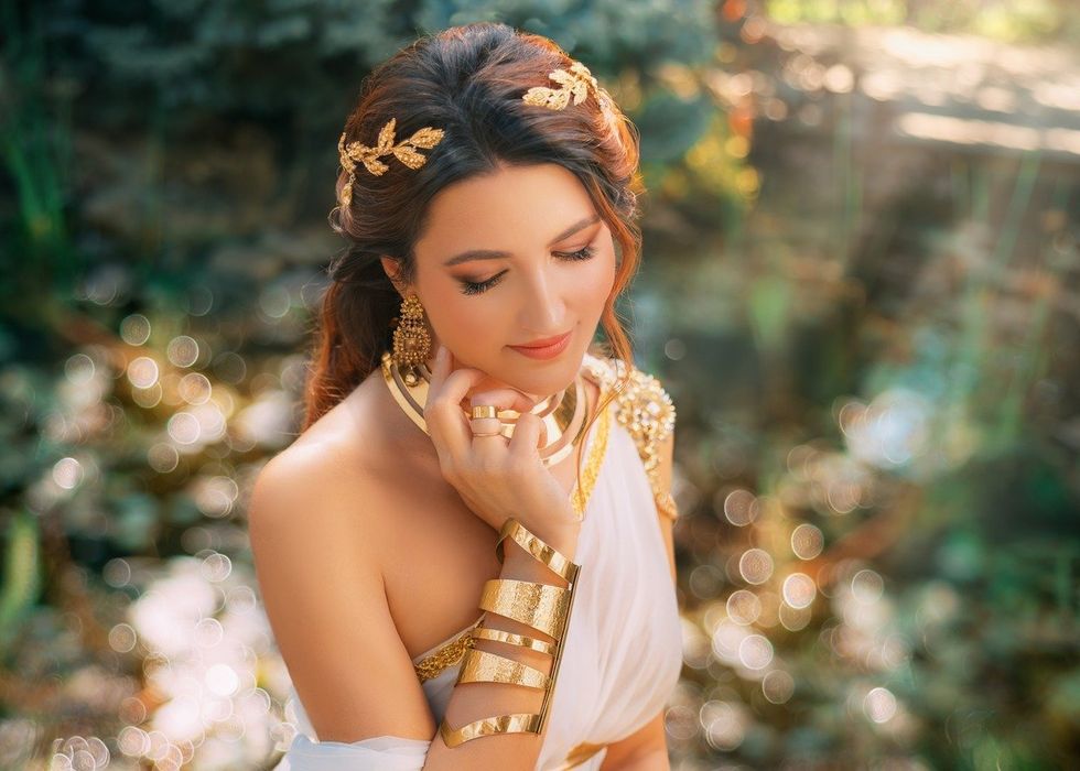 Art photo Fantasy woman greek goddess sits in spring garden green tree grass nature day magic sun light. Sexy girl queen antique style white silk dress, gold jewelry set necklace bracelet crown belt.