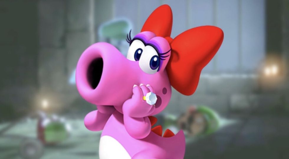Birdo from the Mario franchise. She is a pink dinosaur-esque creature with a large sucker like snout. She has long black eyelashes, an oversized red bow on the back of her head, red scales down her spine and tail, and a white spot on her stomach. She is wearing a large diamond ring on her middle finger.