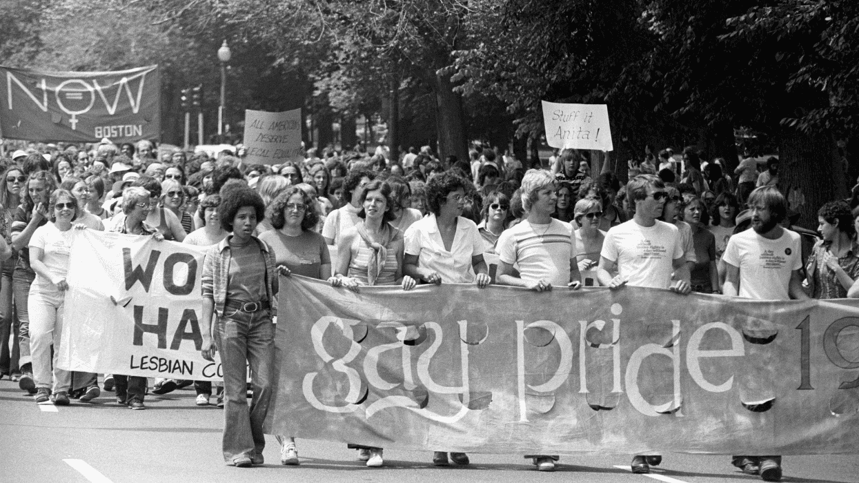 Black-and-white photograph of a large crowd marching in a 1975 Boston Pride parade, with participants carrying banners and signs, including a large “gay pride” banner.