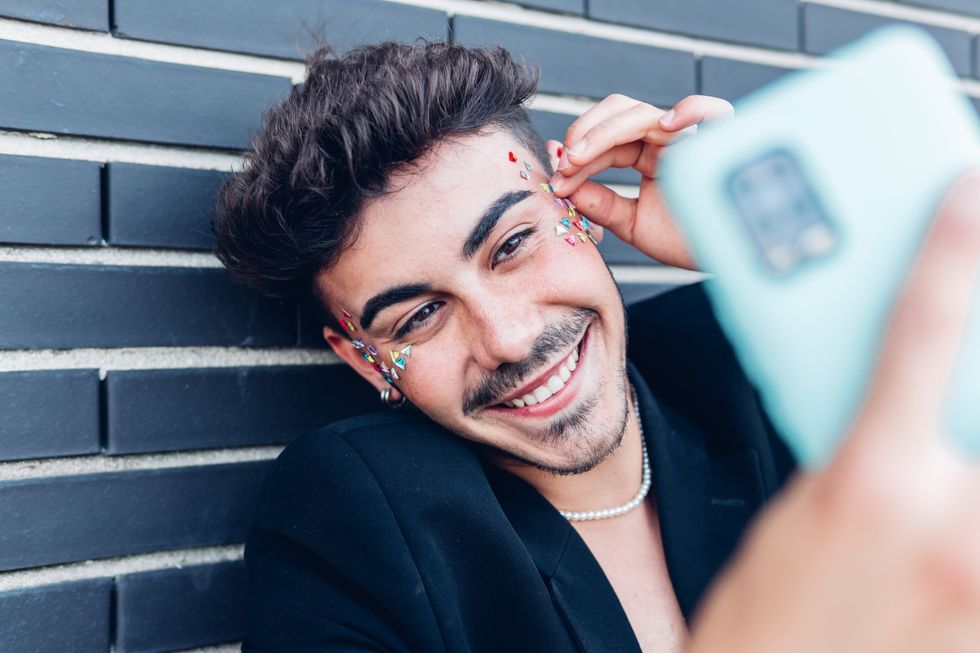 Cheerful young gay man with colorful stickers on face taking self portrait on mobile phone against wall