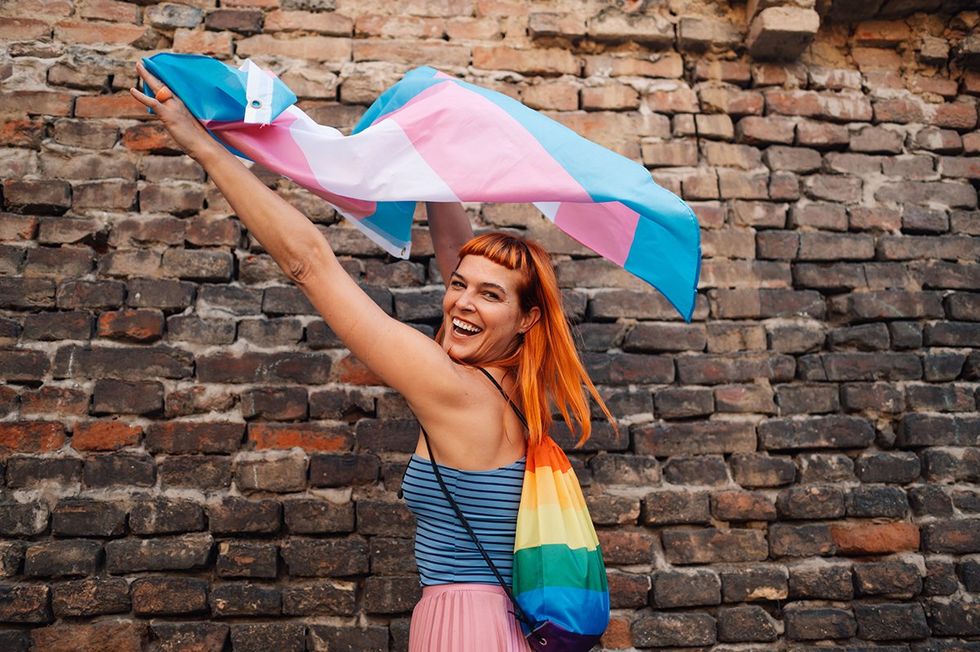 Cheerful young woman with red hair waves a transgender pride flag in front of a brick wall, embodying lgbtq spirit and fighting for equality