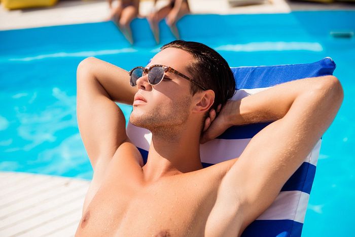 Close up portrait of serene young handsome brunet mature guy in tredy sun glasses, tanning near the pool with arms behind the head, with closed eyes
