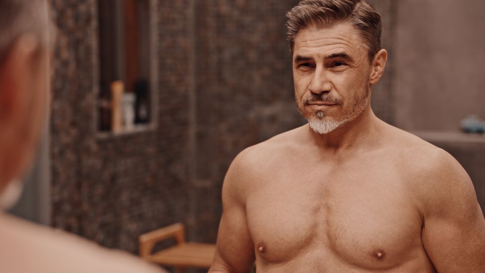 Confident mature male standing shirtless in front of a bathroom mirror. Portrait of muscular fit aged man, smiling.