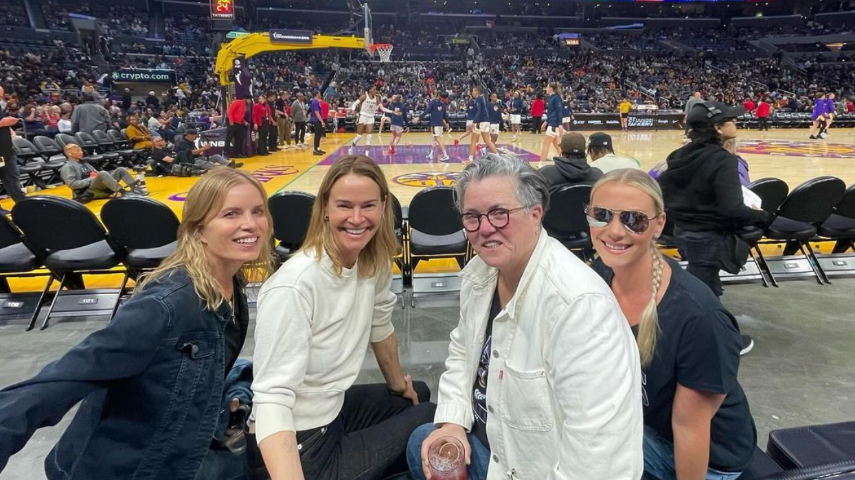 Four white women sitting in second row floor seats at the Los Angeles Sparks vs. Indiana Fever basketball game at the Crypto.com arena. They are, from left to right: Kim Dickens, a 58-year-old with shoulder length blonde hair and a medium-wash denim jacket, Leisha Hailey, a 52-year-old with shoulder length blonde hair, a white sweatshirt, and black jeans, Rosie O'Donnell, a 62-year-old with short, pushed back, silver hair, round, dark rimmed glasses, a white chore coat style jacket, and blue jeans, and Vivienne Rose O'Donnell, a 21-year-old with chest length blonde hair that's been put into a braid and sits over her left shoulder, reflective aviator sunglasses, a black t-shirt and blue jeans.