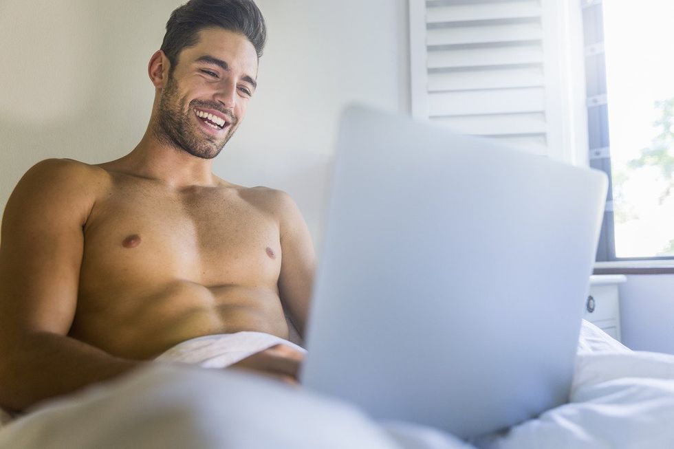 handsome shirtless man in bed smiles at laptop screen