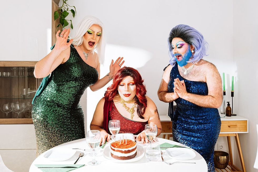 Joyful eccentric male drag queens in wigs and festive dresses with bright makeup screaming happily while standing near friend blowing candles on cake during birthday celebration