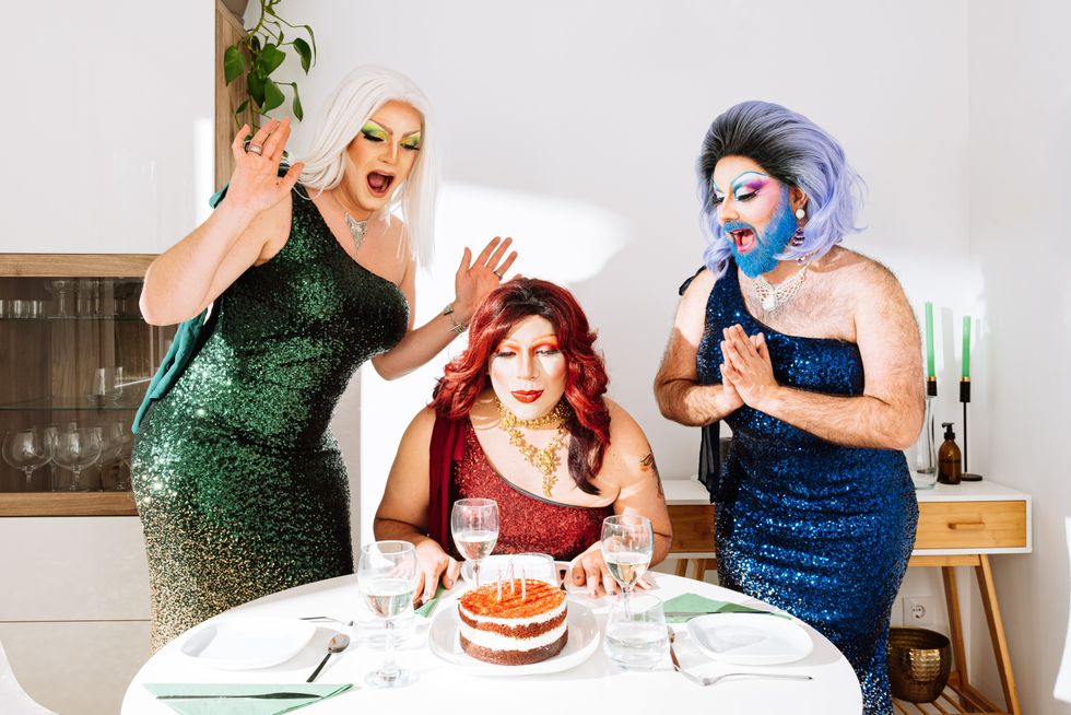 Joyful eccentric male drag queens in wigs and festive dresses with bright makeup screaming happily while standing near friend blowing candles on cake during birthday celebration