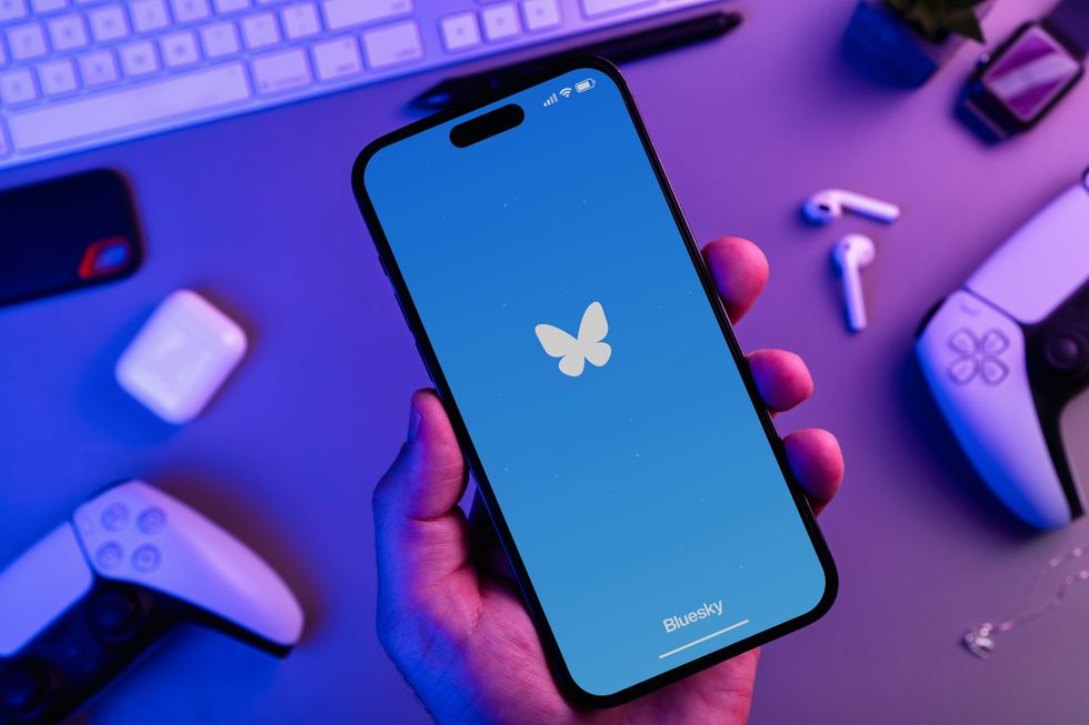 Man holding an iPhone 14 Pro smartphone with Bluesky social media app with PS5 video game controllers. Top view. Office environment. Low blue light. Rio de Janeiro, RJ, Brazil. September 01 2024.