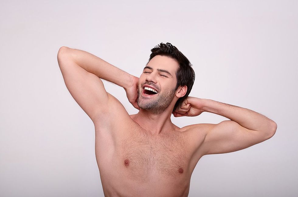 Perfect skin. Portrait of charming topless guy is standing against light background and demonstrating his smooth armpits. He is raising his arms up while looking at camera confidently.