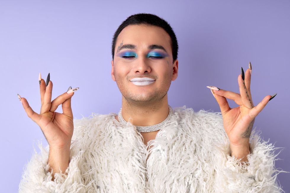 Portrait of fashionable guy in white fluffy coat meditating, engaged in yoga, zen. Womanlike man with bright make-up on face, stand with eyes closed, posing isolated over purple studio background