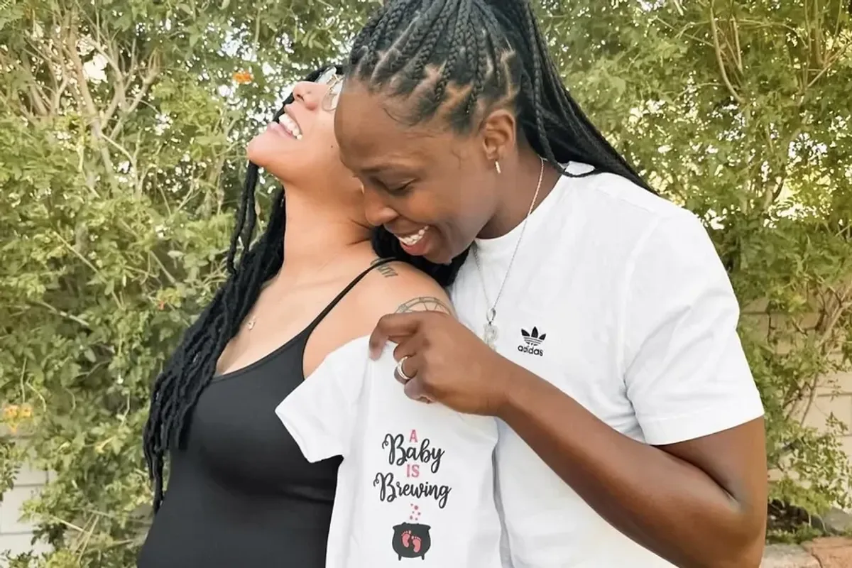 Two Black women with braids smiling, one is pregnant and the other is holding a onesie