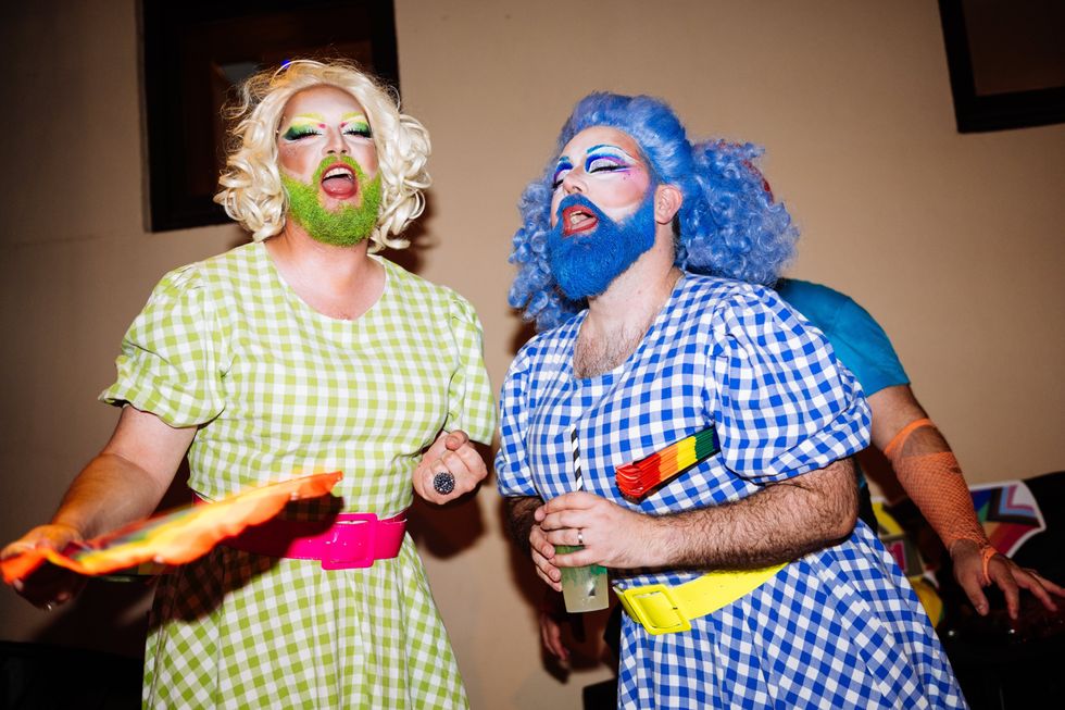 Two drag queens express excitement at a lively queer party, dressed in colorful outfits and sharing a moment of joy. They embody the essence of celebration and inclusivity in the evening atmosphere.