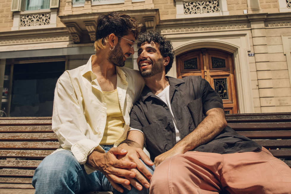 Two men sitting on a bench together, holding hands and staring at one another joyfully.