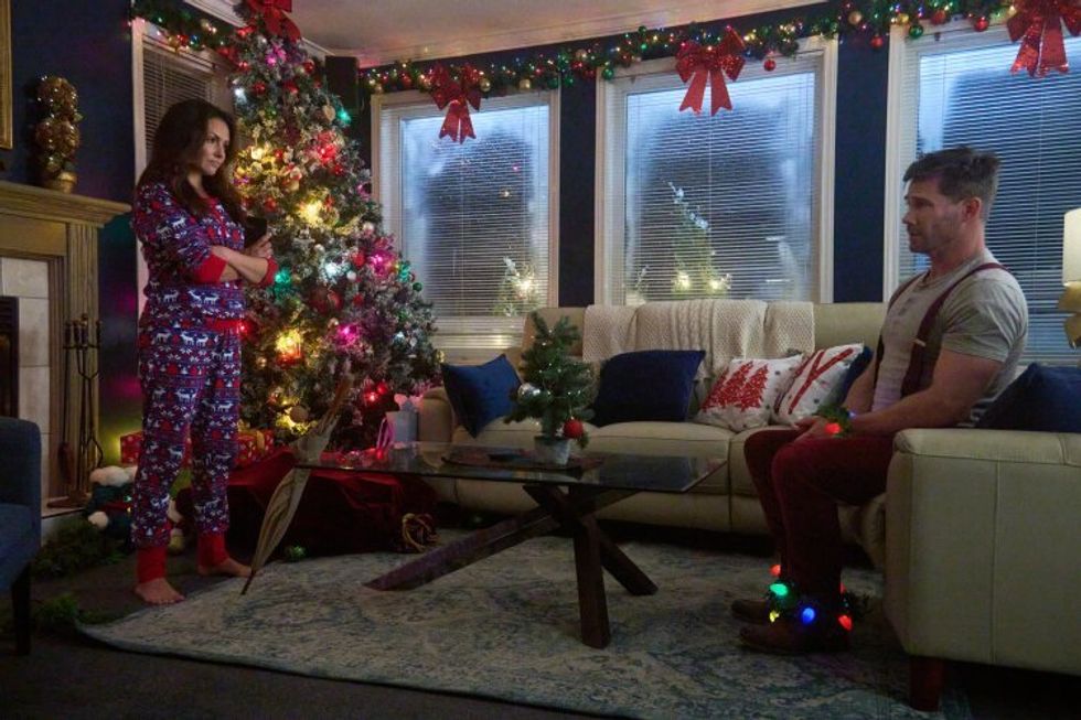 Two people in a living room, one is on the couch tied up with Christmas tree lights