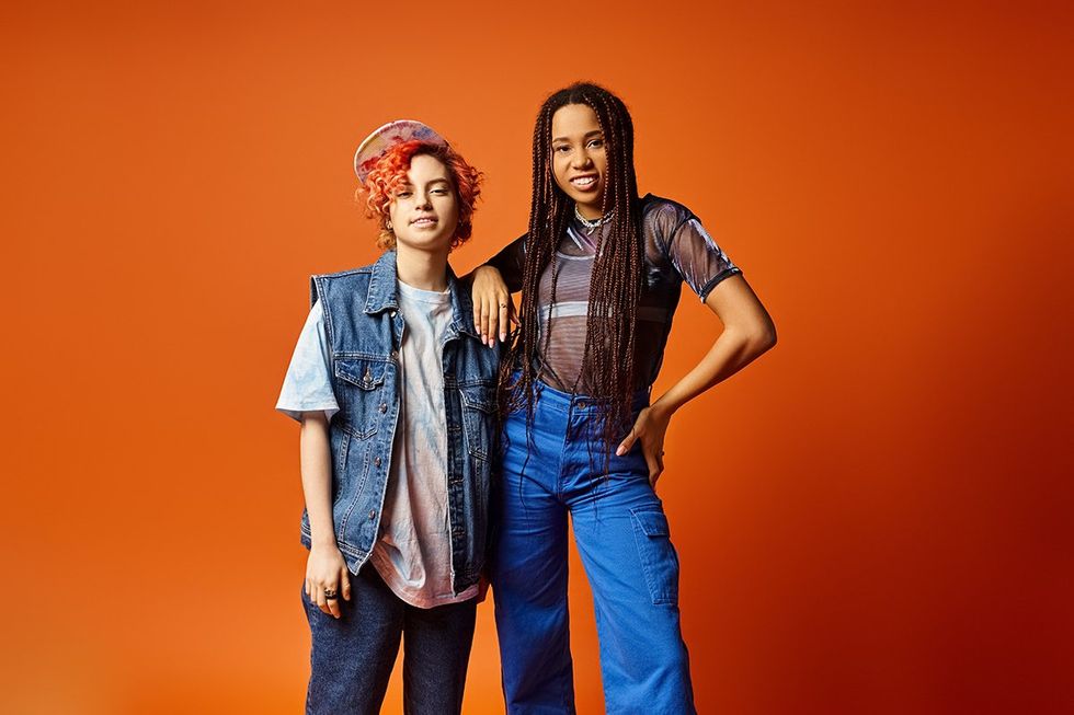 Two stylishly dressed young multicultural friends, including a nonbinary person, standing confidently next to each other in front of a bold orange backdrop.
