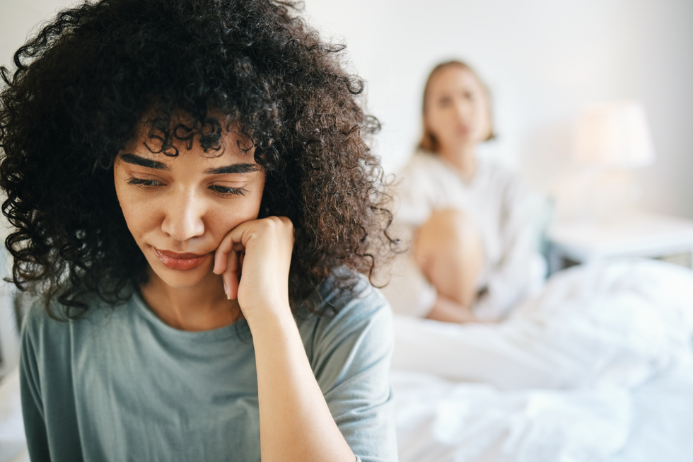 Two women in a bedroom. One, in the back and out of focus, sits at the head of the bed staring at the woman in focus. The woman in focus sits with her head in her hand, looking forlorn.