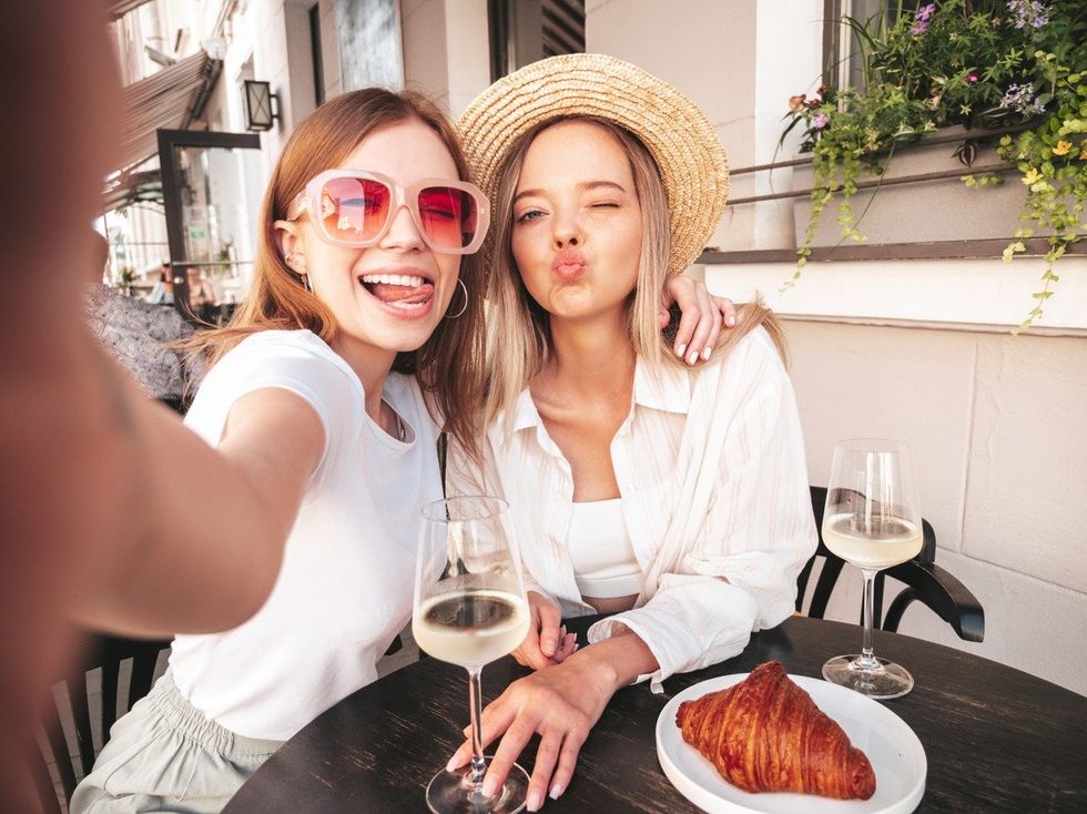 Two young beautiful smiling hipster female in trendy summer clothes.Carefree women posing at veranda cafe in the street.Positive models drinking white wine.Eating croissant.Taking Pov selfie photos