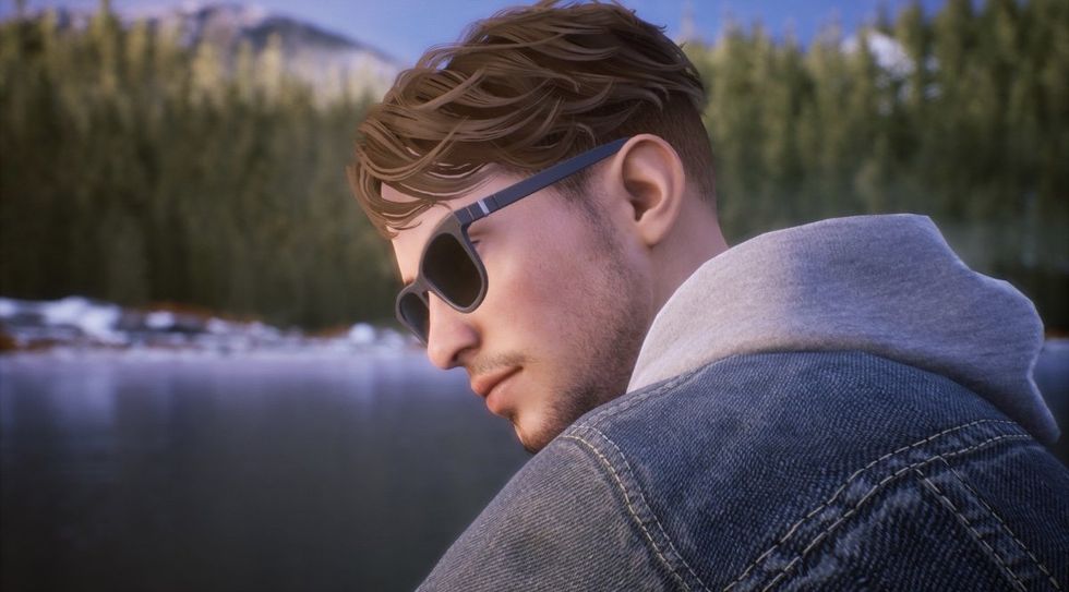 Tyler Ronan from the video game Tell Me Why. He is a 21-year-old white trans man with short light brown hair and a stubbly beard. He is sat by a lake, looking over his shoulder, wearing black sun glasses and a jean jacket over a heather grey hoodie.